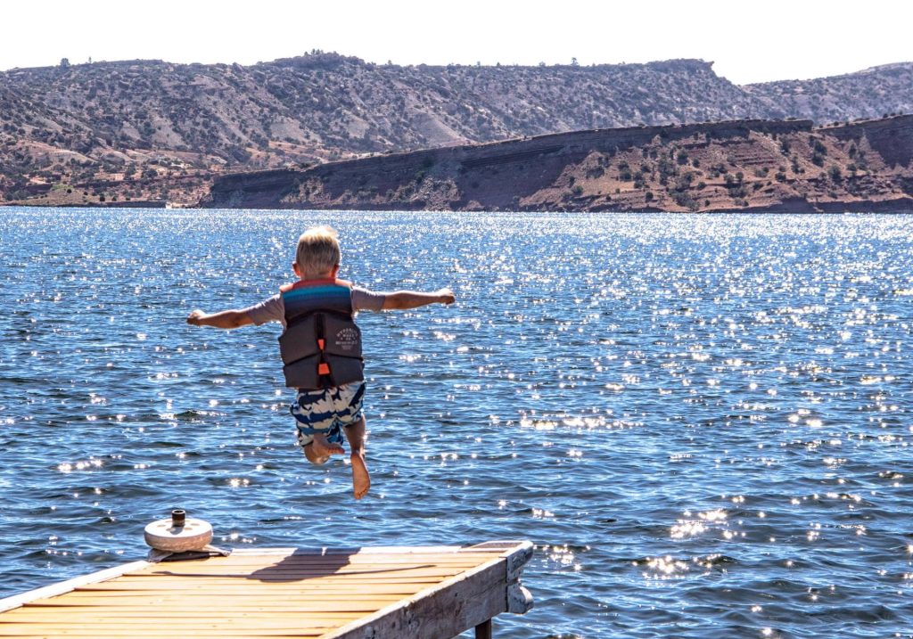 A boy jumps off a dock into the blue waters of a lake with vegetated hills in the background
