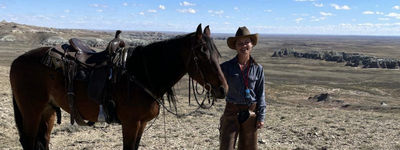 Gabby stands beside a horse in a sagebrush landscape