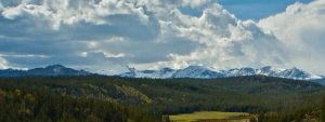 Green, forested hills in middleground, with snow-covered mountains rising above beneath a clouded sky.