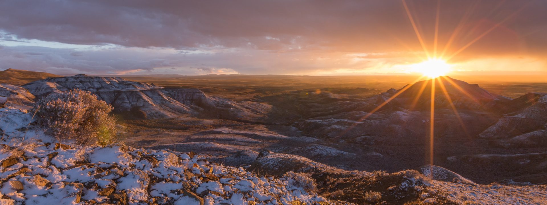 View of the rising sun from the rim of an overlook covered with snow-dusted sagebrush.